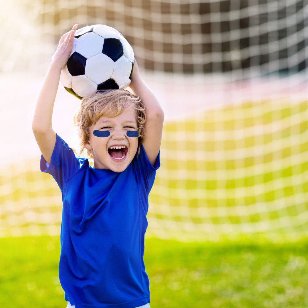 A young boy in a blue soccer jersey stands on a grassy field in front of a soccer goal, holding a black and white soccer ball above his head with both arms raised. He has blue face paint on his eyes.