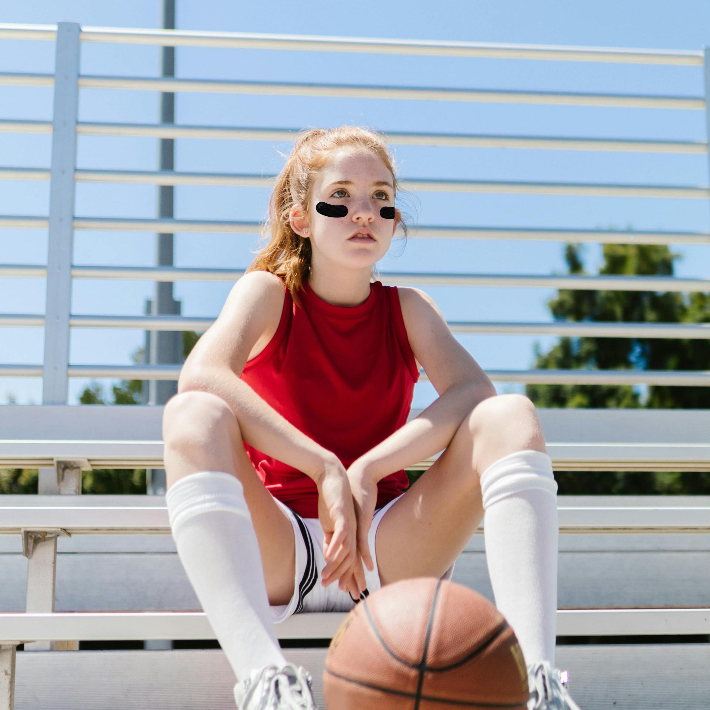 A young woman in a red sleeveless athletic top, white knee-high socks, and white sneakers sits on outdoor bleachers with a basketball in front of her. She is wearing black eye masks, and the background shows a blue sky and metal railing.