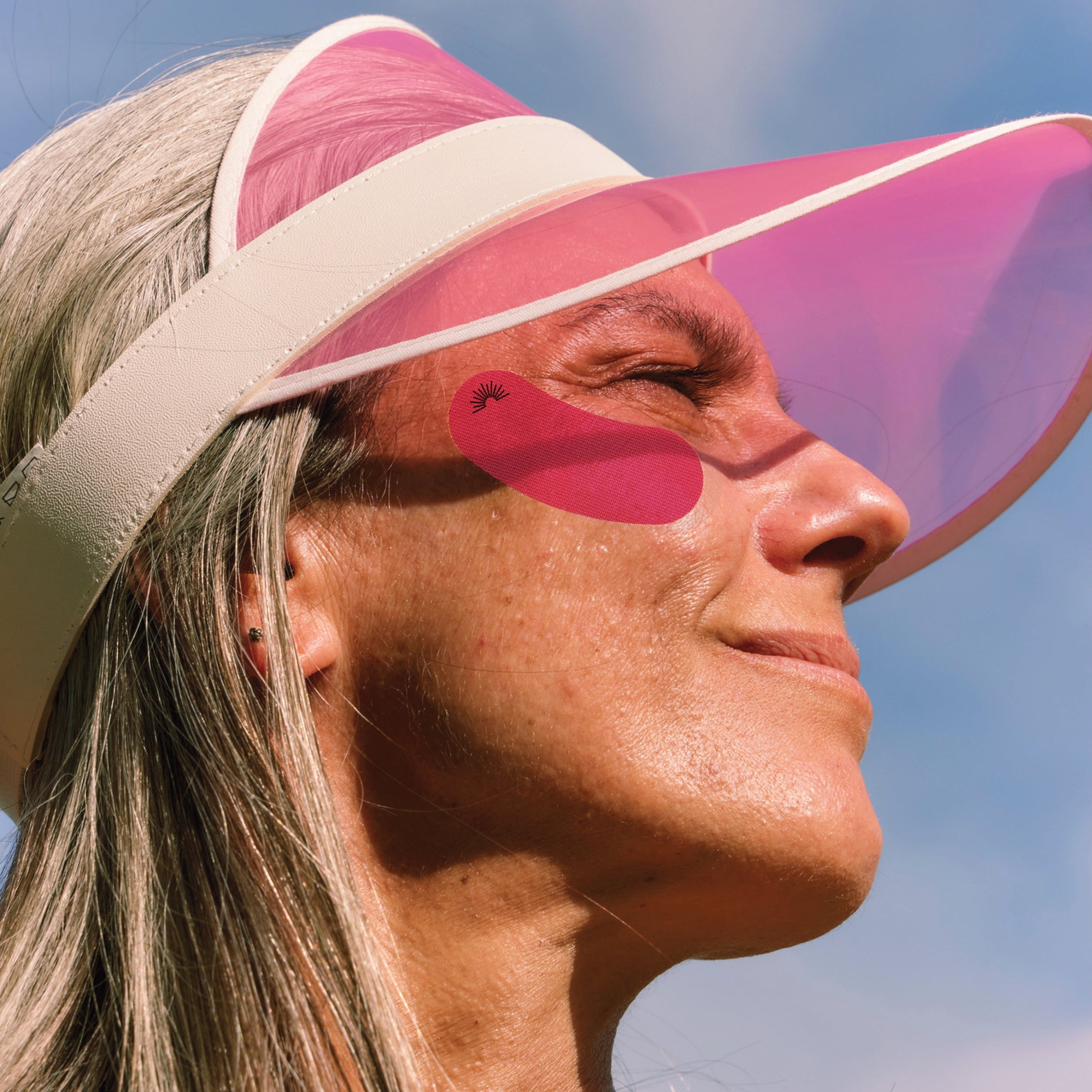 A close-up lifestyle shot of a woman outdoors wearing a pink visor with a white headband and a pink face shield, against a blue sky.