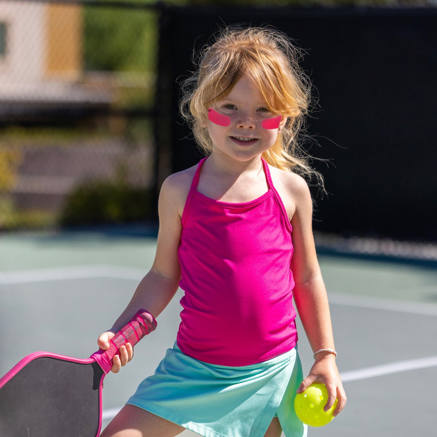 A young girl with blonde hair stands on an outdoor pickleball court wearing a bright pink tank top, a light blue skirt, and pink sunglasses, holding a pink paddle and a yellow pickleball.