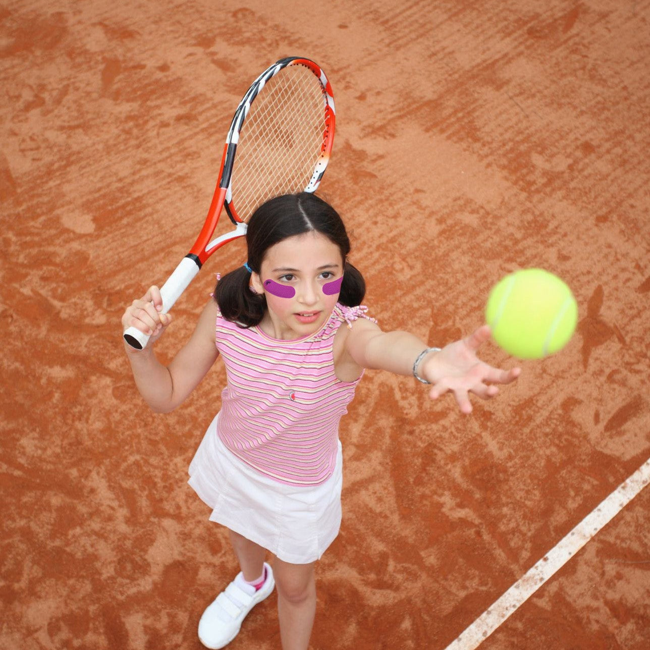 A young girl with dark hair in pigtails stands on a clay tennis court holding a tennis racket and reaching toward a yellow tennis ball. She is wearing a pink and white striped top, a white skirt, and white sneakers.